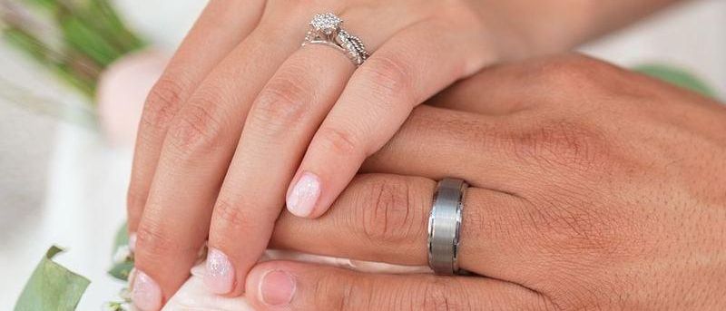 Ein Mann und eine Frau halten Händchen, beide tragen Ringe. Der Ring der Frau hat einen Diamanten. Sie stehen nah beieinander, möglicherweise auf einer Hochzeit.