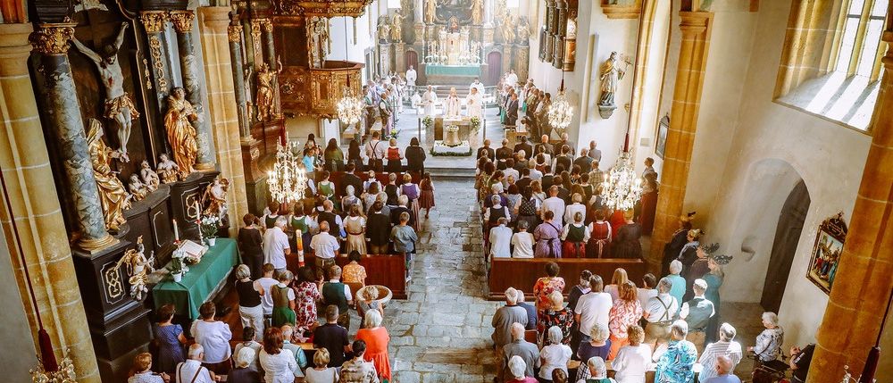 Eine große Gemeinde besucht einen Gottesdienst in einer Kirche mit verzierten Säulen, Kronleuchtern und einem zentralen Altar, der mit Blumen geschmückt ist.