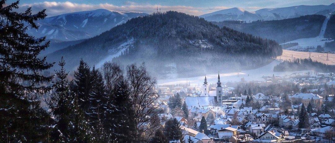 Eine winterliche Landschaft mit einem verschneiten Dorf unter einem Bergrücken, bedeckt mit Schnee, mit einer Kirche und einem Turm im Vordergrund, unter einem blauen Himmel mit Wolken.