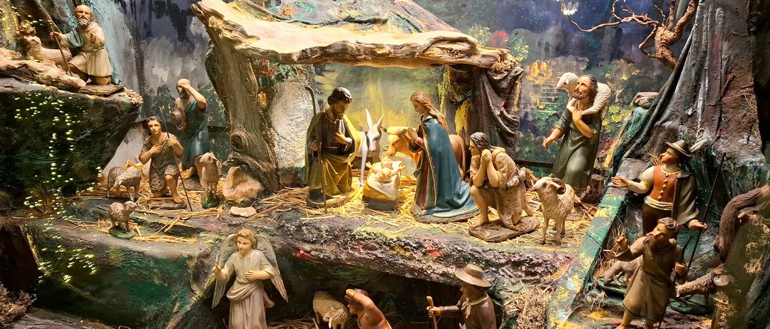 Eine Krippe mit Maria, Josef und Jesus in der Krippe, umgeben von anderen Figuren, einschließlich Hirten, Tieren und einem Engel. Sie befindet sich in einer Höhle mit einem Steindach.