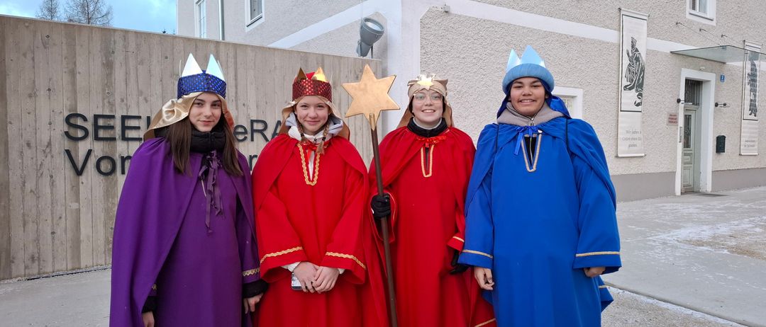 Vier Personen in aufwendigen Kostümen und Kronen posieren für ein Foto vor einem Gebäude mit Fenstern. Die Frau in der Mitte hält einen Stern.