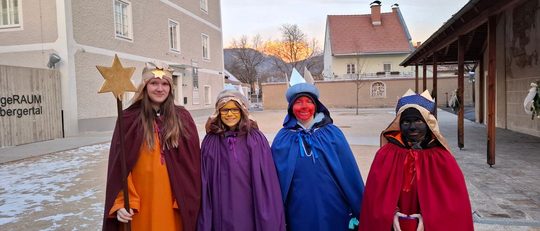 Vier Personen in farbenfrohen Kostümen mit Gesichtsbemalung stehen vor einem Gebäude mit Bergen im Hintergrund.