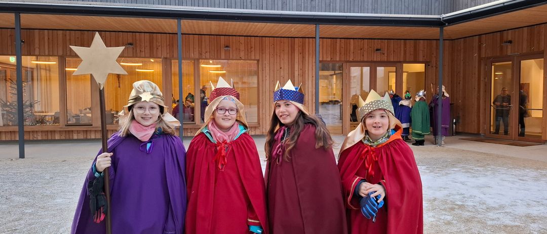 Vier Mädchen in roten Umhängen und Kronen stehen vor einem hölzernen Gebäude. Sie lächeln und posieren für ein Foto.