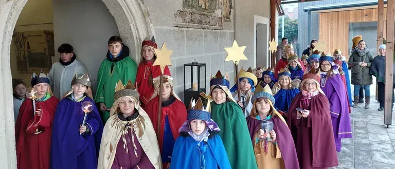 Eine Gruppe von Kindern in bunten Kostümen mit Kronen und Sternen steht in einer Reihe, möglicherweise für ein Weihnachtsspiel oder eine Parade. Sie halten Requisiten und stehen vor einem Gebäude.
