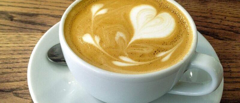 Eine Tasse Kaffee mit herzförmiger Schaumkreation steht auf einem Unterteller auf einem Holztisch.