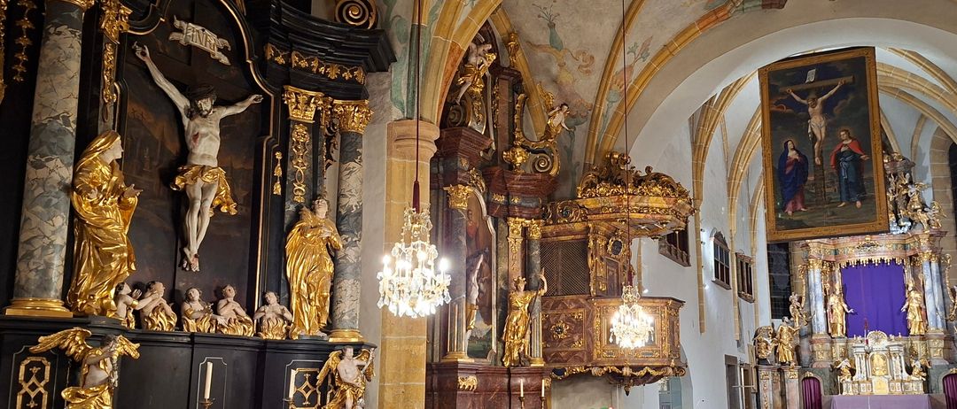 Das Innere einer Kirche zeigt kunstvolle Holzbänke, einen goldenen Altar und ein Kruzifixgemälde. Kronleuchter hängen von der Decke, und Statuen schmücken die Wände.