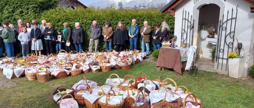 Eine Gruppe von Menschen steht vor einer Kapelle mit vielen dekorierten Körben auf dem Boden. Einige Personen halten Blumen, und in der Nähe steht ein Tisch mit einer Kerze.