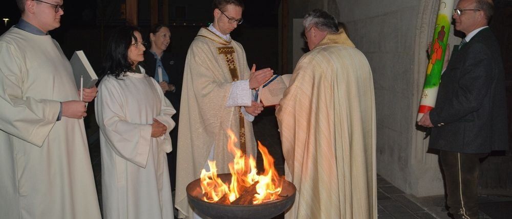 Zwei Priester stehen vor einem Feuerschale, einer liest ein Buch und der andere hält eine Kerze. Zwei Frauen stehen hinter ihnen.