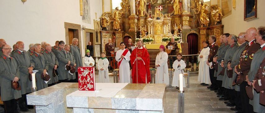 Eine Gruppe von Menschen in Uniformen und Gewändern steht um einen Altar in einer Kirche. Sie halten Kerzen und schauen nach vorne, wo ein Priester eine Zeremonie leitet.