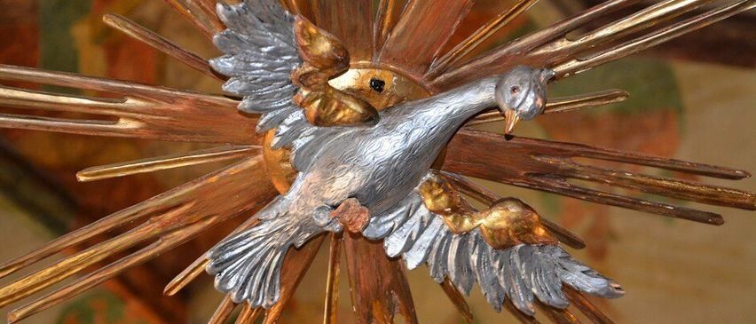 Eine Nahaufnahme einer metallischen Taube mit ausgebreiteten Flügeln, zentral auf einem Sonnenstrahl mit goldenen Strahlen positioniert. Die Taube hat einen glänzenden silbernen Körper und goldene Akzente an ihren Flügeln und ihrem Schnabel.