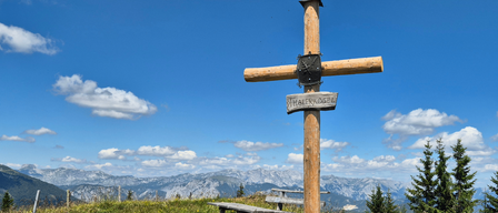 Ein Holzschild mit der Aufschrift 'Berggottesdienst am Thalerkogel' steht auf einer Wiese mit einer Bank und einer Bergkette im Hintergrund. Das Datum und die Uhrzeit der Veranstaltung sind ebenfalls angezeigt.