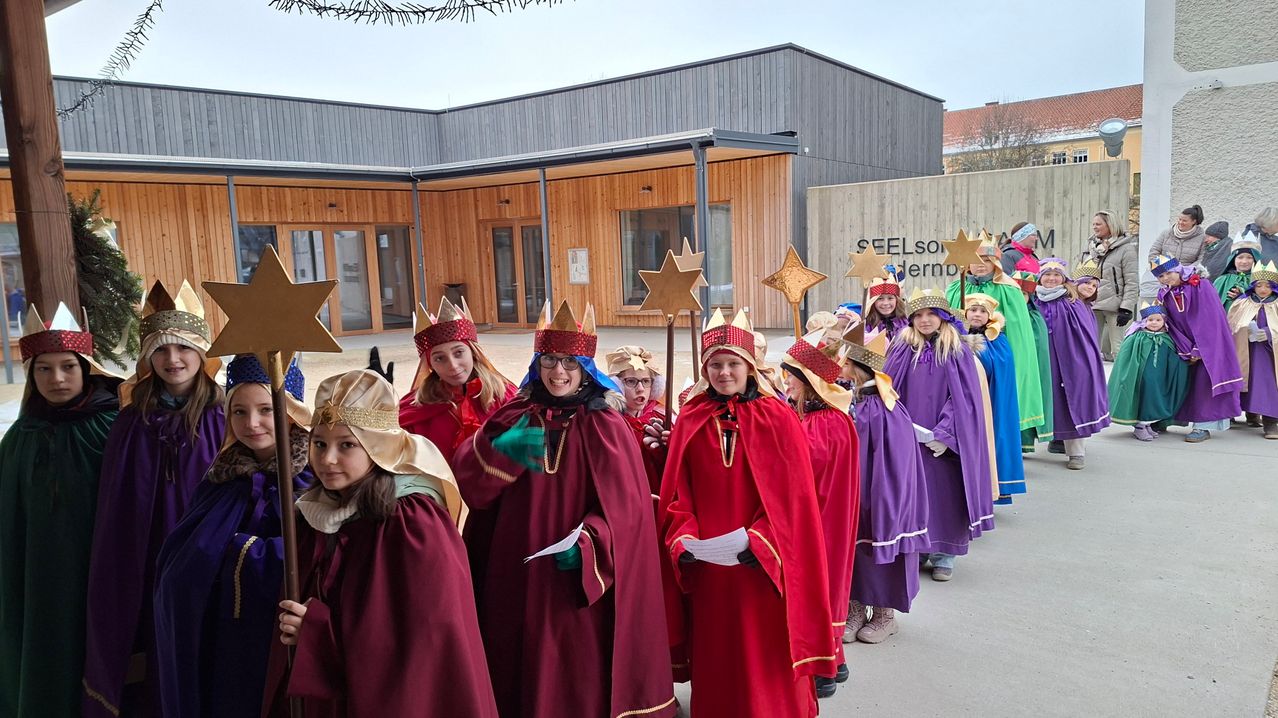 Eine Gruppe von Kindern, verkleidet als Weise, hält Sterne in der Hand und steht vor einem Gebäude. Sie tragen Kronen und rote Umhänge.