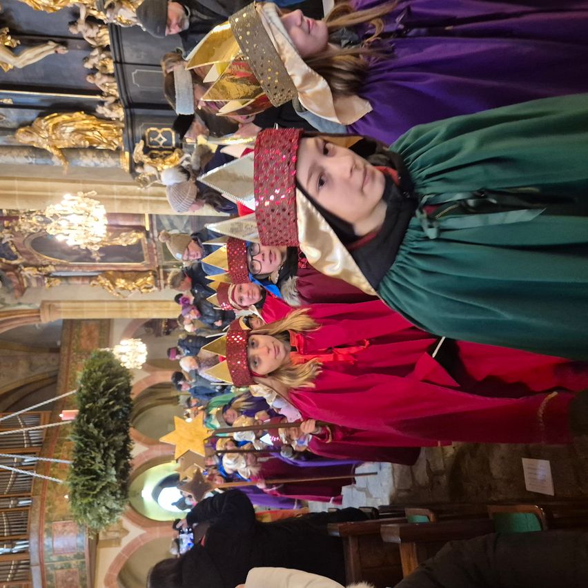 Eine Gruppe von Kindern in aufwendigen Kostümen, einschließlich Kronen und Umhängen, versammelt sich in einer Kirche. Sie scheinen an einer festlichen Veranstaltung teilzunehmen, möglicherweise einer Weihnachtsfeier.