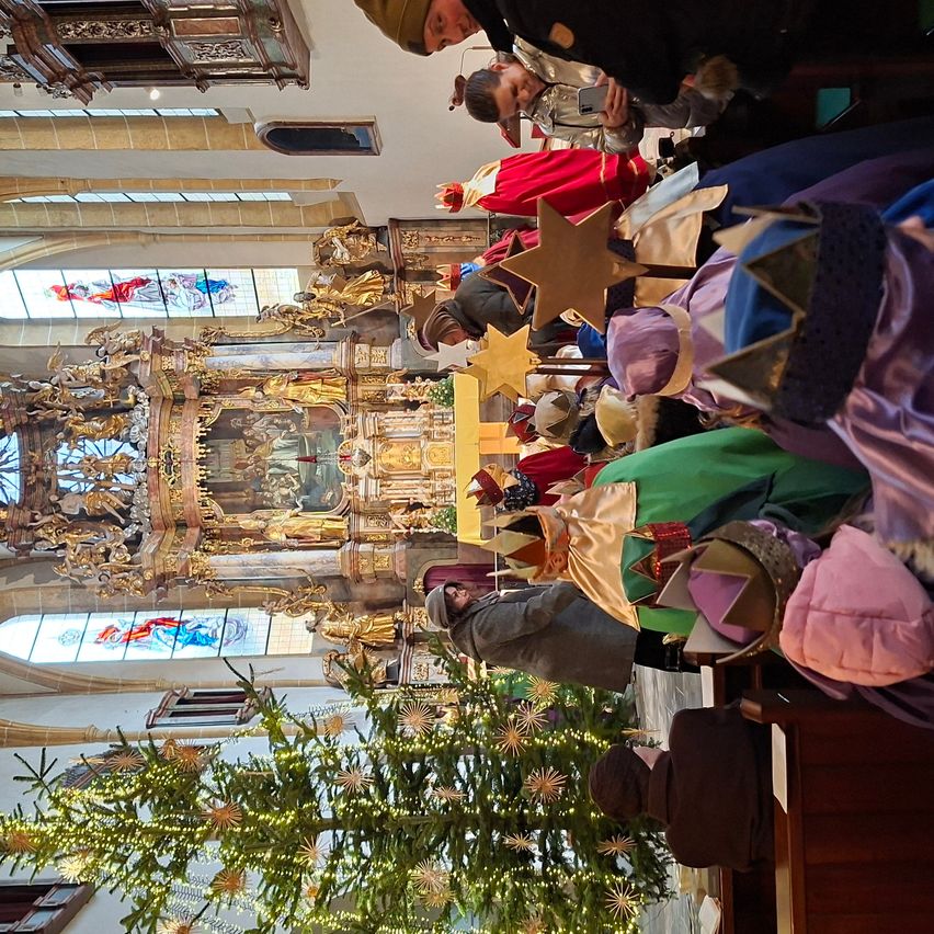 Auf einem Kirchenaltar versammeln sich mehrere Personen in Kostümen und Kronen um einen geschmückten Weihnachtsbaum. Ein Mann macht ein Foto.