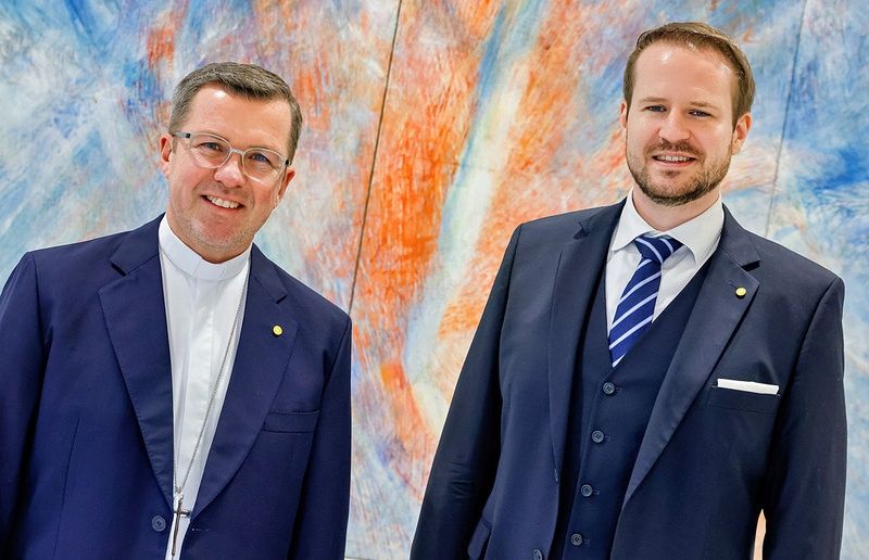 Two men in suits, one with a visible cross necklace, smile at the camera against a vibrant abstract backdrop.