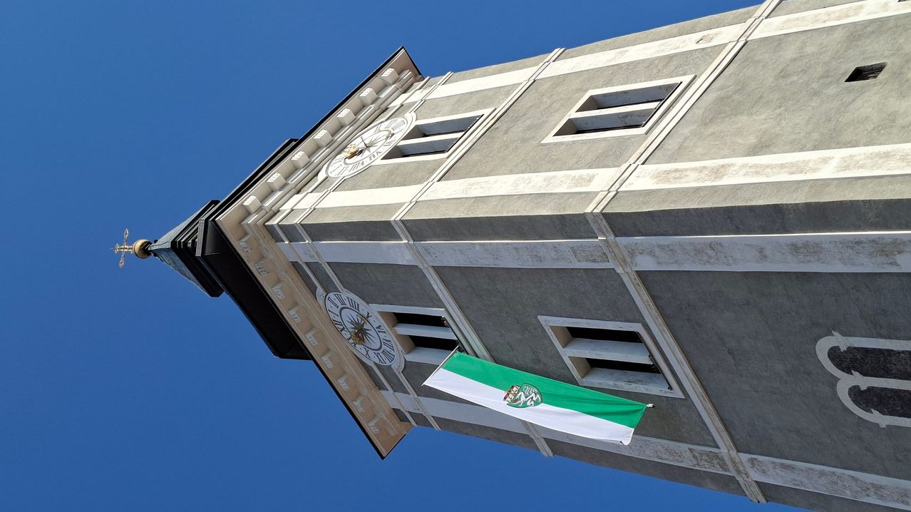 Ein Uhrturm mit einer Fahne unter den Uhren. Die Fahne ist grün und weiß mit einem Logo in der Mitte. Der Uhrturm ist aus Beton.
