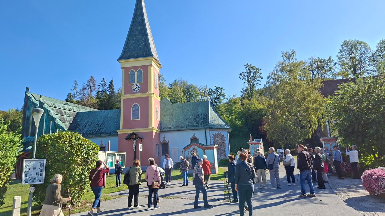 Eine Gruppe von Menschen besucht eine Kirche mit einem Turm, einer Uhr und einem Kreuz an der Wand. Sie stehen auf dem Weg vor der Kirche.