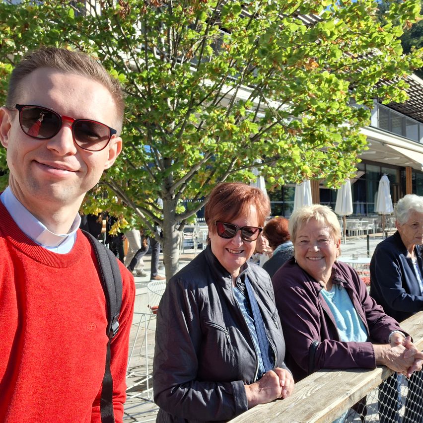 Ein Mann in einem roten Pullover und Sonnenbrille steht mit zwei älteren Frauen, eine in einem lila Jacke und die andere in einem blauen Shirt, vor einem Baum und einem Gebäude.