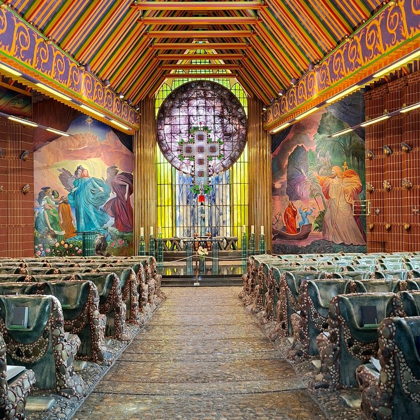 Eine farbenfrohe Kapelle mit bunten Glasfenstern, leuchtenden Wandgemälden und einzigartigen Kirchenbänken. Der zentrale Altar zeigt ein großes Kreuz, und die Wände sind mit kunstvollen Mustern verziert.