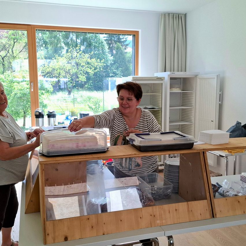Zwei Frauen stehen bei einem Tisch mit Essen in einer Küche. Eine Frau hält einen Kuchendeckel. Ein Kühlschrank und ein Fenster mit Bäumen draußen sind im Hintergrund.