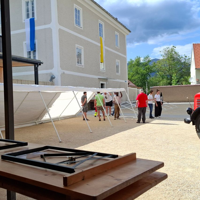 Mehrere Personen stehen vor einem Gebäude mit einem Zelt und Fahnen. Ein rotes Fahrzeug ist in der Nähe geparkt.