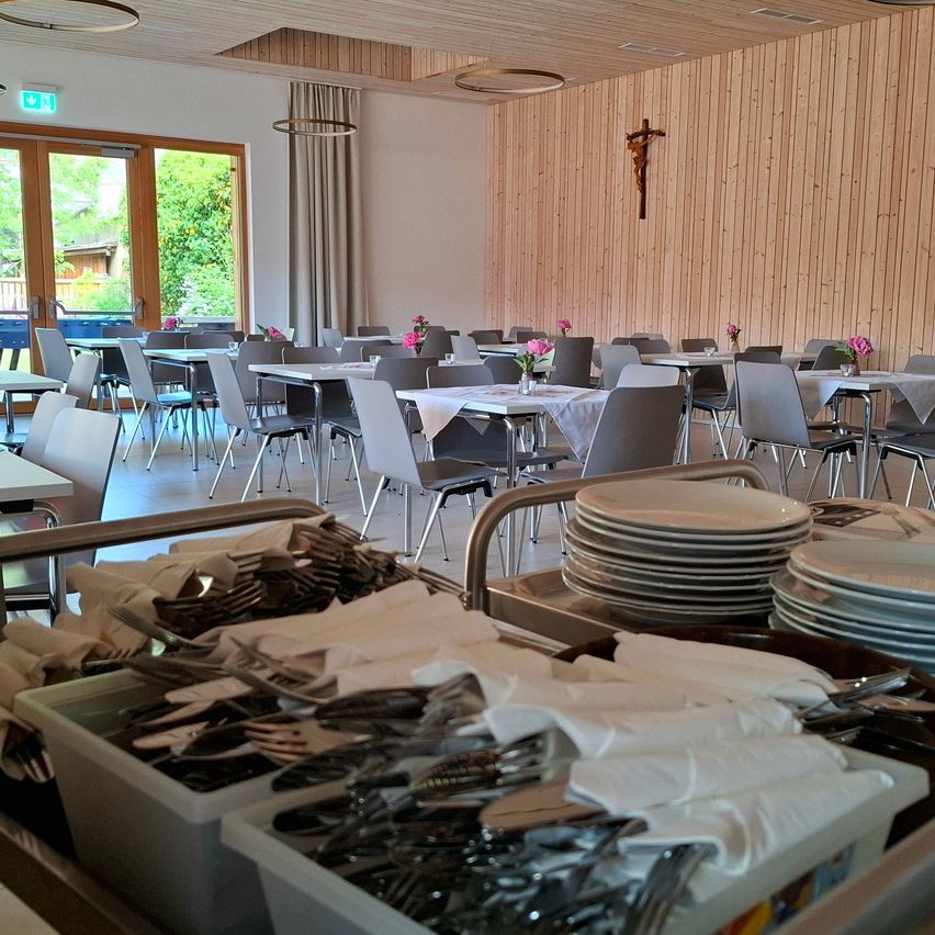 Ein Innenrestaurant mit Holzwänden hat viele leere Tische und Stühle. Die Tische sind mit weißen Tischdecken bedeckt und haben Blumen in der Mitte. Ein Stapel Teller steht vor dem Tisch. Ein Kreuz ist an der Wand montiert.