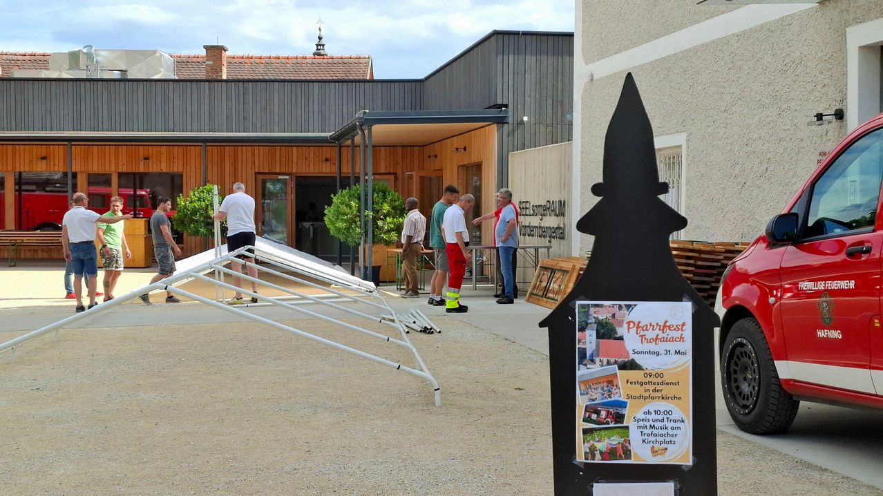 Mehrere Männer stehen vor einem Gebäude. Einer hält eine Metallstruktur. Ein Schild auf einem Pfosten steht 'Pfarrfest Trofaiach'.