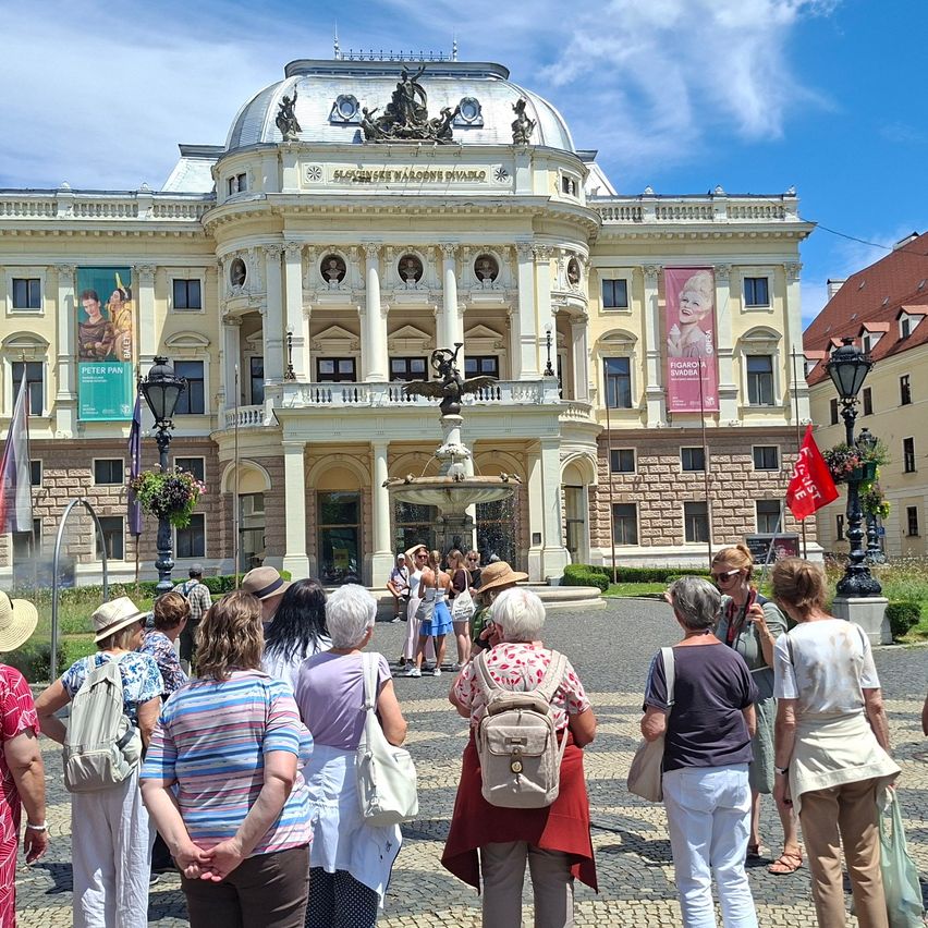 Tourists gather outside a historic theater with a fountain, banners, and statues. The building has classical architecture.