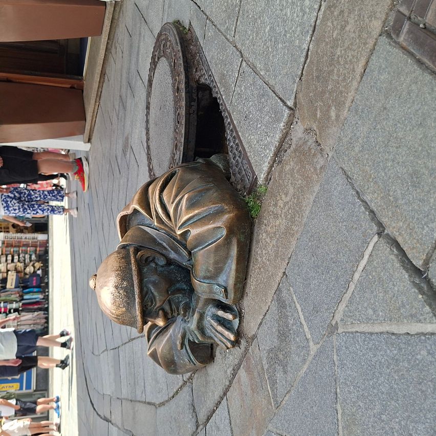 A bronze statue of a sleeping man is embedded in the sidewalk near a shop with items on display. Several people walk by.