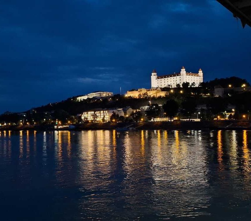 A castle illuminated at night stands on a hill above a river, with city lights reflecting on the water.