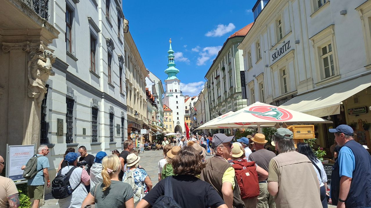 A crowded street with tourists in front of a clock tower. Buildings line the street, some with signs and flags. The sky is clear with passing clouds.