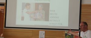 Ein Mann hält eine Präsentation mit einer Projektionsleinwand hinter ihm, die ein Bild von zwei Personen und den Text 'Impulsvortrag' zeigt.