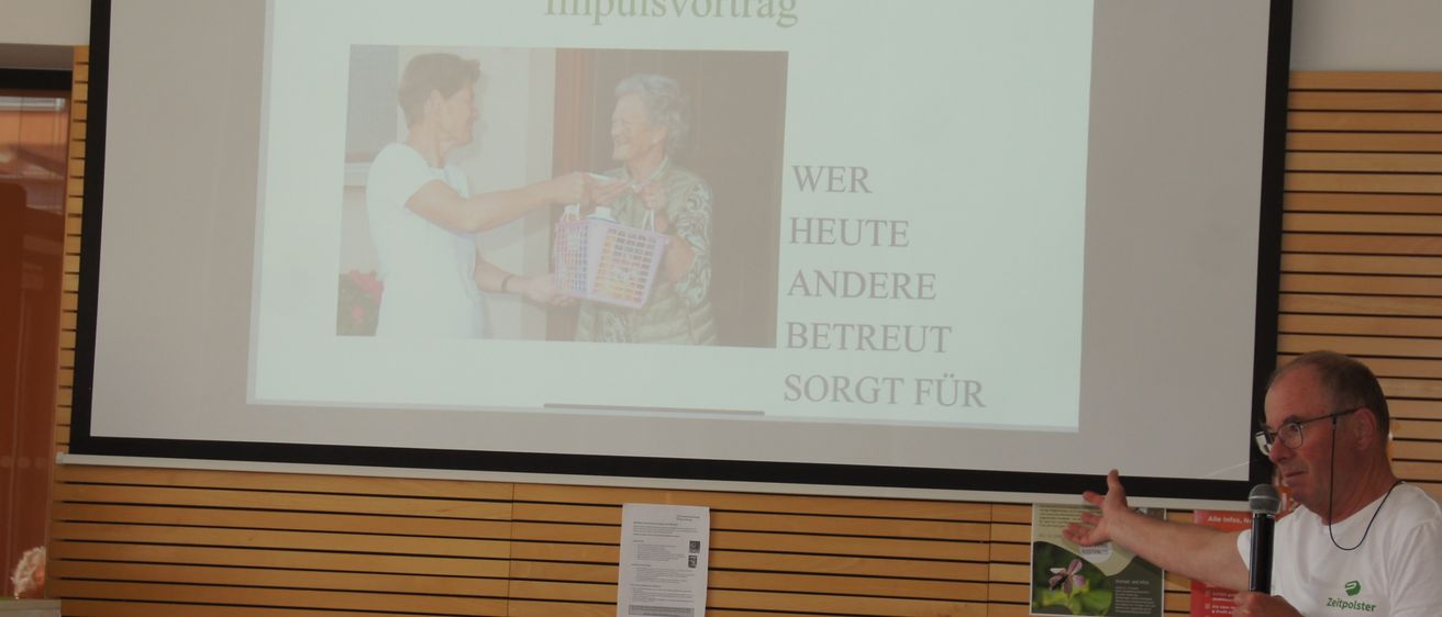 Ein Mann hält eine Präsentation mit einer Projektionsleinwand hinter ihm, die ein Bild von zwei Personen und den Text 'Impulsvortrag' zeigt.