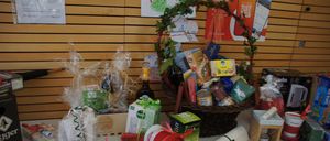 Eine Ausstellung verschiedener Geschenkkörbe und Artikel auf einem Tisch, einschließlich eines großen geflochtenen Korbes voller Produkte, verpackte Boxen, Handtücher und Flaschen. Der Hintergrund zeigt eine Holzwand mit Postern.