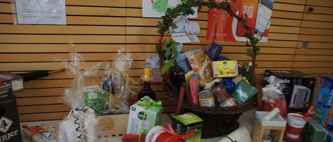 Eine Ausstellung verschiedener Geschenkkörbe und Artikel auf einem Tisch, einschließlich eines großen geflochtenen Korbes voller Produkte, verpackte Boxen, Handtücher und Flaschen. Der Hintergrund zeigt eine Holzwand mit Postern.