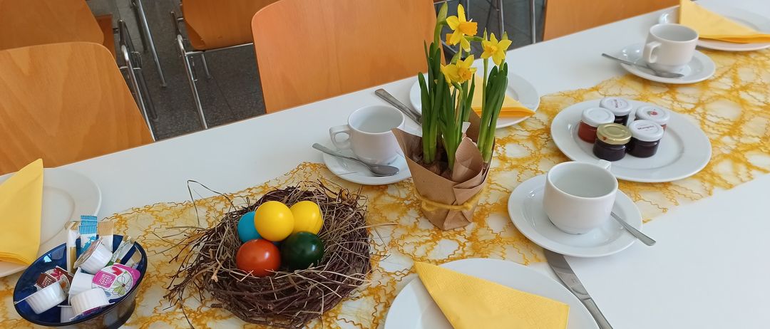 Ein Tisch ist mit einer Schüssel Ostereier, einem gelben Blumenzentrum, Tassen, Untertassen, Löffeln und Servietten gedeckt. Im Hintergrund sind Stühle.