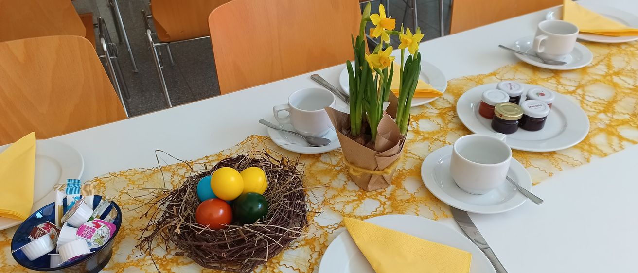 Ein Tisch ist mit einer Schüssel Ostereier, einem gelben Blumenzentrum, Tassen, Untertassen, Löffeln und Servietten gedeckt. Im Hintergrund sind Stühle.