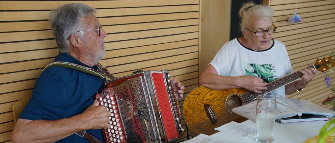 Ein älterer Mann spielt Akkordeon, während eine ältere Frau Gitarre spielt. Sie sitzen an einem Tisch mit Notenblättern und einem Mikrofon.
