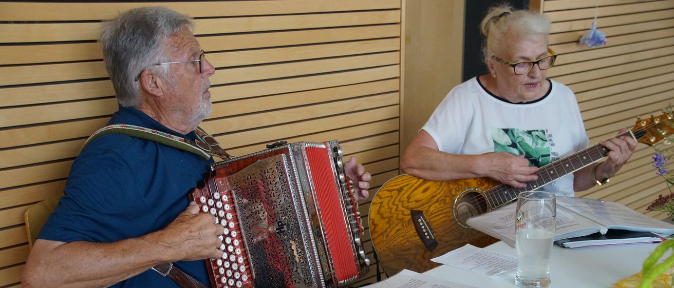 Ein älterer Mann spielt Akkordeon, während eine ältere Frau Gitarre spielt. Sie sitzen an einem Tisch mit Notenblättern und einem Mikrofon.