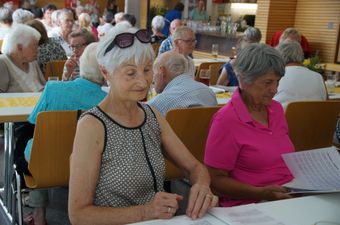 Eine ältere Frau mit Sonnenbrille auf dem Kopf sitzt mit einer anderen Frau am Tisch und schaut beide auf Papiere vor sich. Sie sind von anderen Menschen umgeben, die an Tischen in einem Restaurant sitzen.