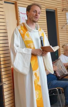 Ein Priester in einer weißen Robe mit einem gelben Schalkragen hält ein Buch und ein Mikrofon und hält möglicherweise eine Rede.