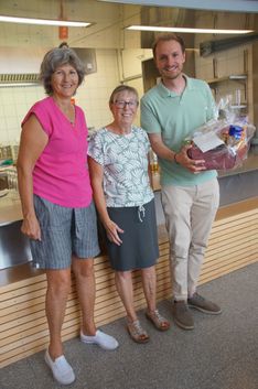 Drei Menschen stehen in einer Küche, zwei Frauen und ein Mann, lächeln und posieren für ein Foto. Der Mann hält einen Geschenkkorb.