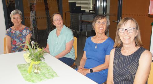 Zwei Frauen sitzen an einem Tisch mit einer Blumenarrangement, lächeln und schauen zur Kamera. Dahinter ist ein Raum mit Stühlen und einem Tisch zu sehen.