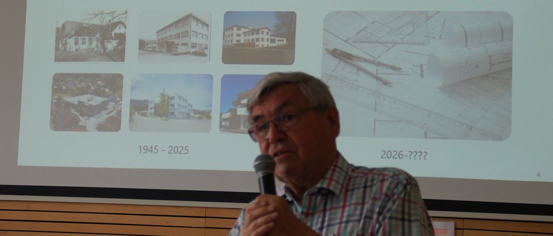 Ein Mann steht vor einer Leinwand und spricht in ein Mikrofon. Die Leinwand zeigt eine Präsentation über die Geschichte von BAUR, beginnend von 1945 bis 2025. Die Präsentation enthält auch Bilder von Gebäuden.