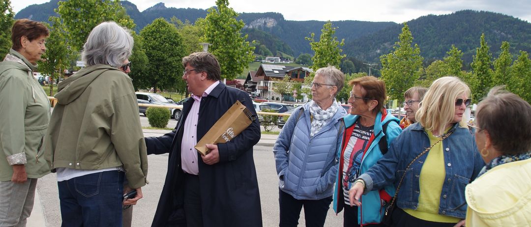Eine Gruppe von Menschen ist in einem Außenbereich versammelt. Ein Mann in der Mitte hält einen Beutel und spricht mit anderen. Sie stehen im Kreis, mit Bergen im Hintergrund.