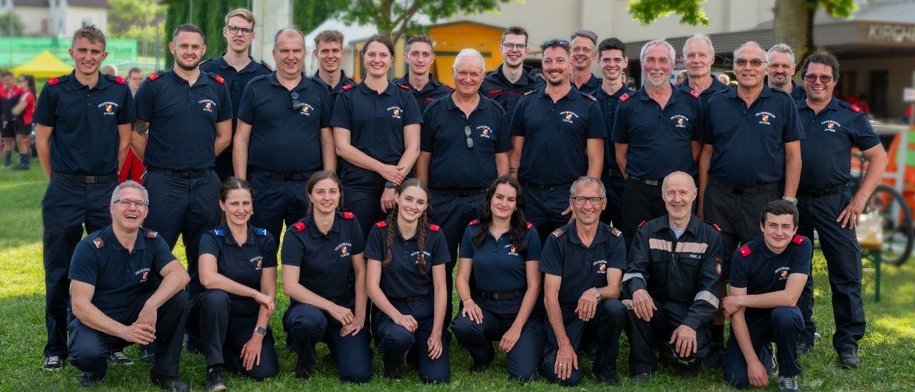 Eine Gruppe uniformierter Personen, wahrscheinlich Feuerwehrleute, posiert für ein Foto auf einer Rasenfläche mit Bäumen und einem Gebäude im Hintergrund.