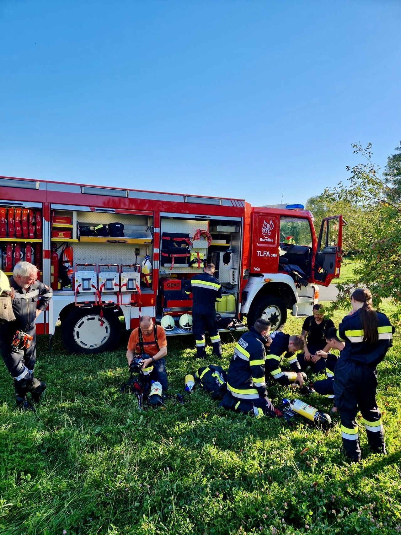 Eine Gruppe von Feuerwehrleuten in Uniform versammelt sich um einen roten Feuerwehrwagen mit offener Tür, einige knien und andere stehen, sie machen wahrscheinlich eine Trainingsübung.