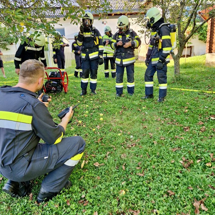 Eine Gruppe von Feuerwehrleuten in voller Ausrüstung versammelt sich auf einer Rasenfläche, einer kniet und hält ein Funkgerät, während andere um ihn herum stehen.