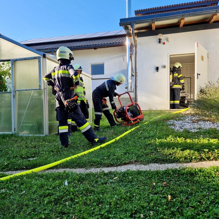 Feuerwehrleute in voller Montur arbeiten vor einem Haus, einer zieht eine rote Maschine mit einem gelben Schlauch.