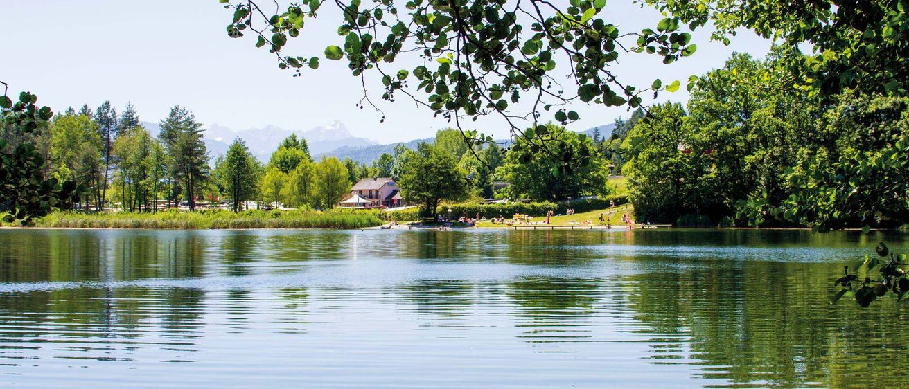 Ein ruhiger See mit einem Haus und Bergen im Hintergrund, umgeben von üppiger Grünfläche. Mehrere Menschen entspannen sich am Ufer.